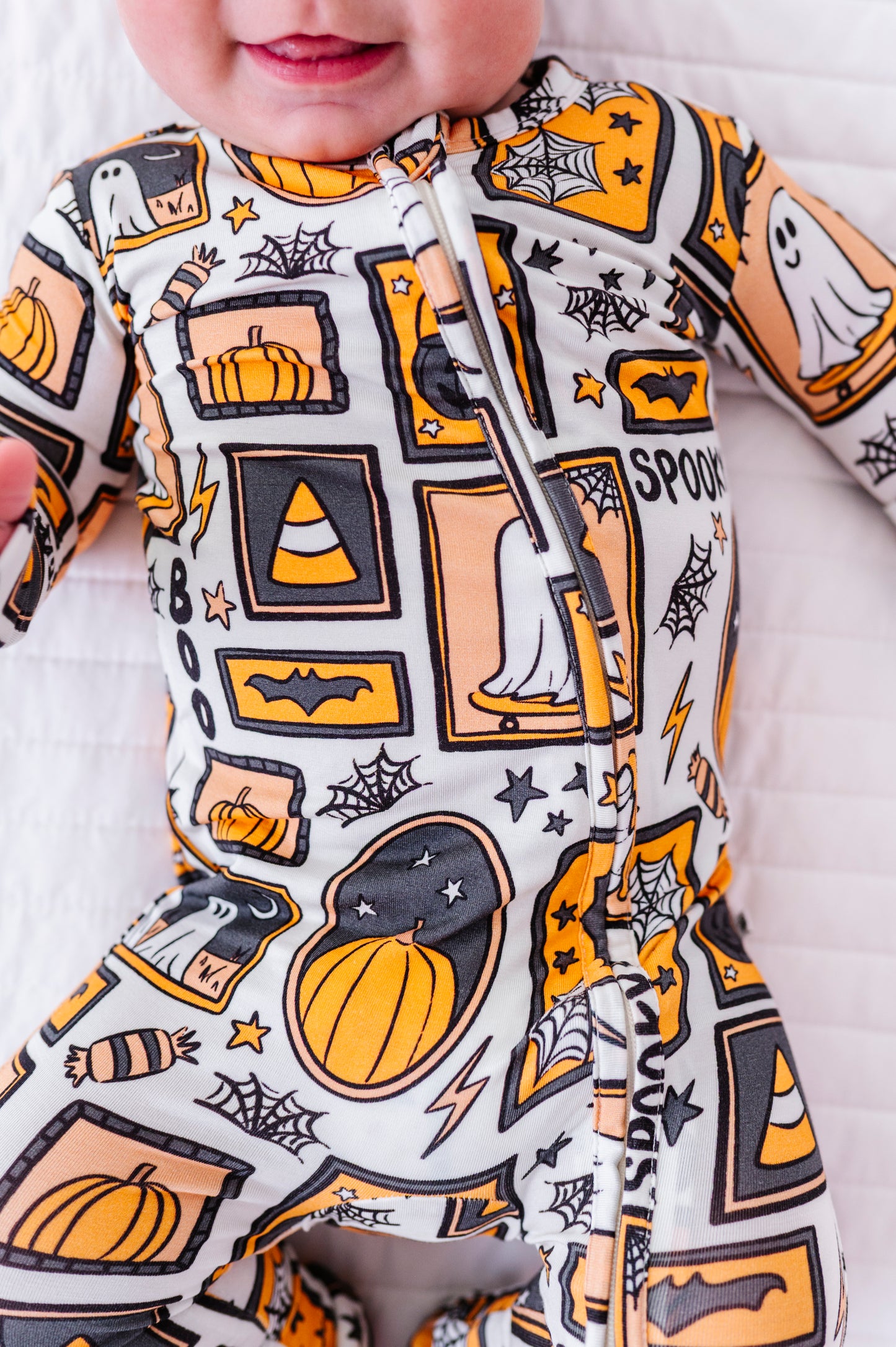 A baby wearing a white one-piece sleeper with a vibrant orange, black, and yellow Halloween pattern featuring pumpkins, ghosts, bats, and spiderwebs.