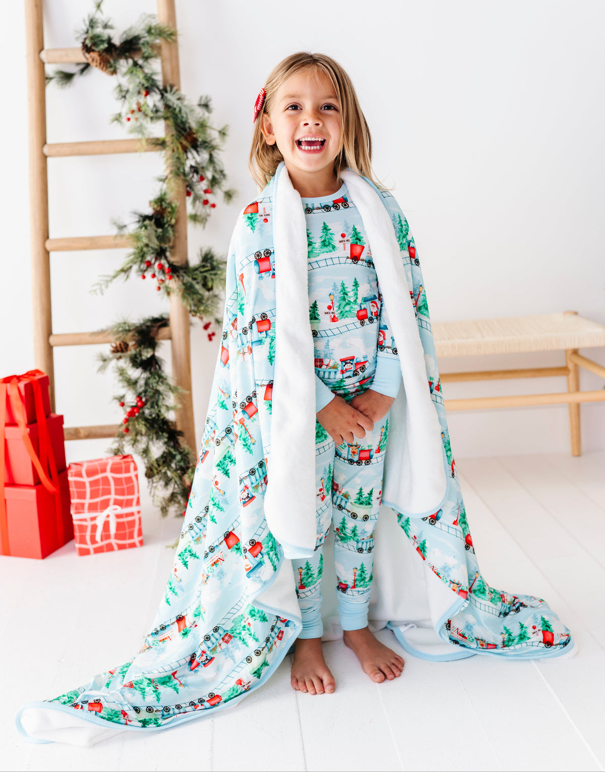 A young girl stands barefoot in a bright white room, wrapped in a light blue and white holiday-themed bamboo blanket with a matching patterned top and pants. She is smiling, with a wooden ladder decorated with greenery and red ornaments behind her, and wrapped in a white blanket.