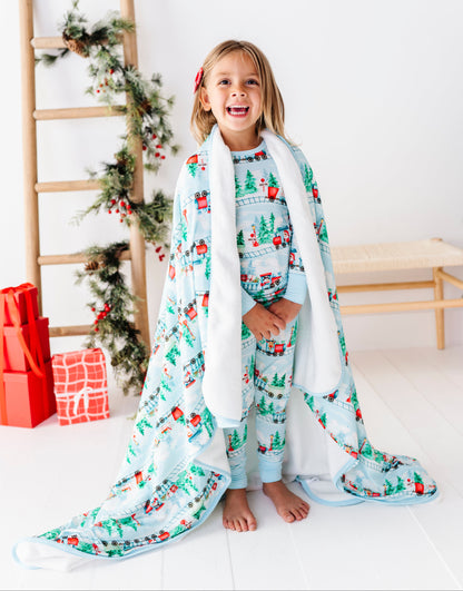 A young girl stands barefoot in a bright white room, wrapped in a light blue and white holiday-themed bamboo blanket with a matching patterned top and pants. She is smiling, with a wooden ladder decorated with greenery and red ornaments behind her, and wrapped in a white blanket.