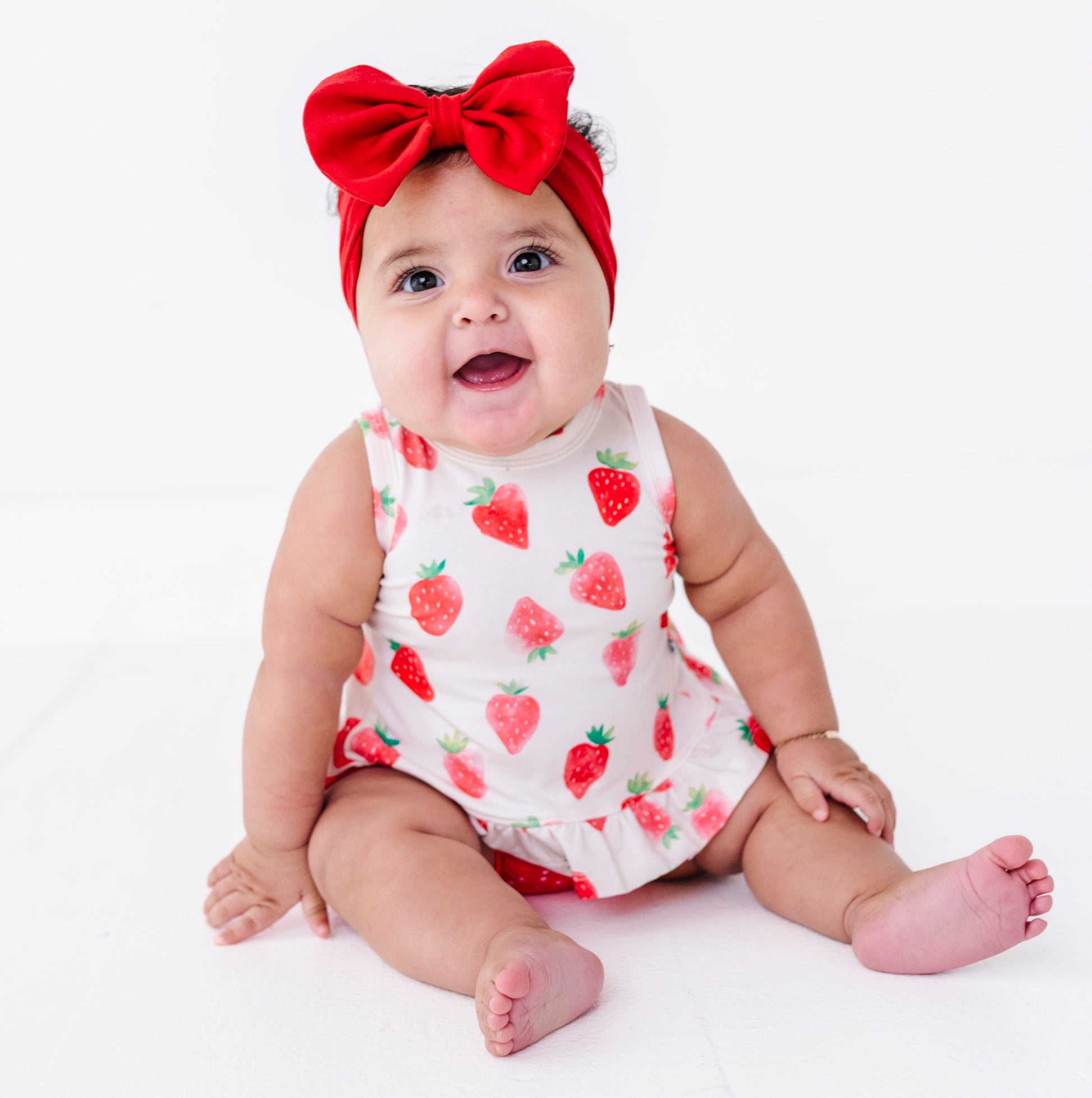 Baby wearing a strawberry-patterned outfit and red headband on a white background