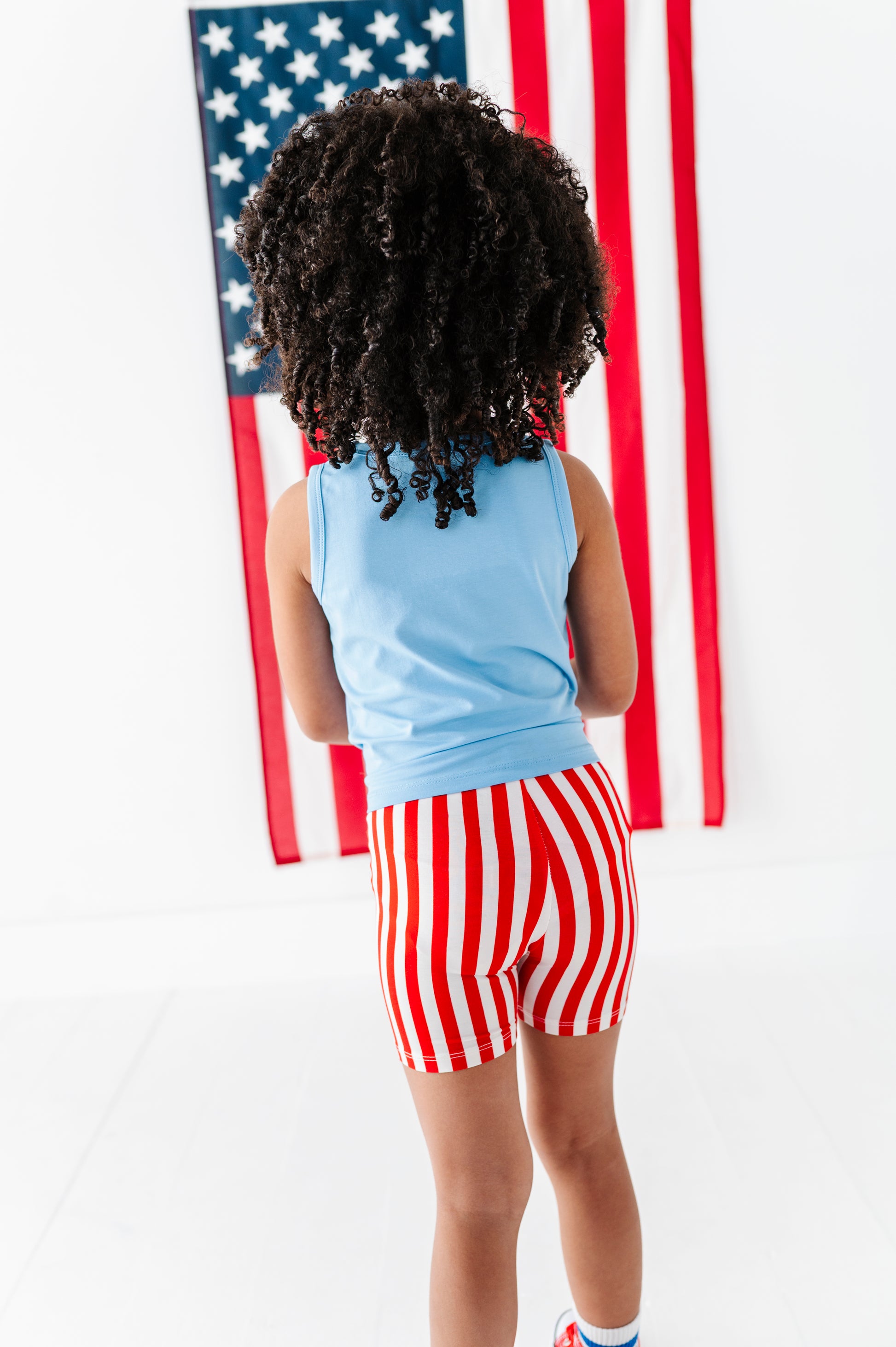 Child wearing a blue bamboo tank top and red and white striped bamboo biker shorts standing in front of an American flag.
