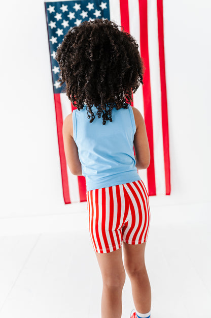 Child wearing a blue bamboo tank top and red and white striped bamboo biker shorts standing in front of an American flag.