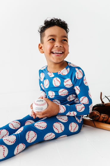 Child wearing a blue bamboo pj shirt and pants with baseball patterns, holding a baseball and smiling.