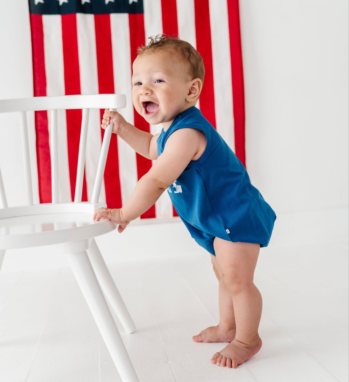 A baby standing on a white floor and leaning on a white chair, wearing a bright blue sleeveless romper. An American flag hangs on the wall behind the baby.