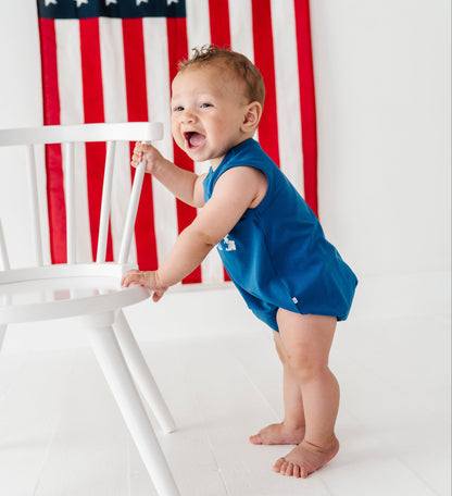 A baby standing on a white floor and leaning on a white chair, wearing a bright blue sleeveless romper. An American flag hangs on the wall behind the baby.
