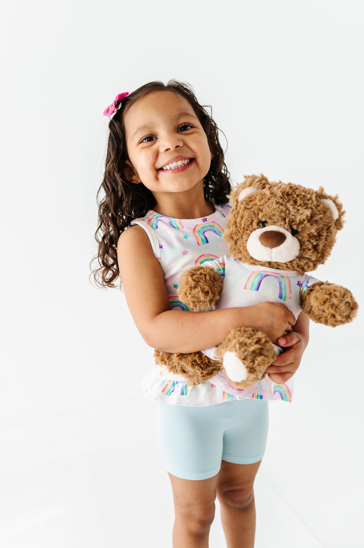 Young girl holding a teddy bear wearing a matching watercolor rainbow outfit against a white background