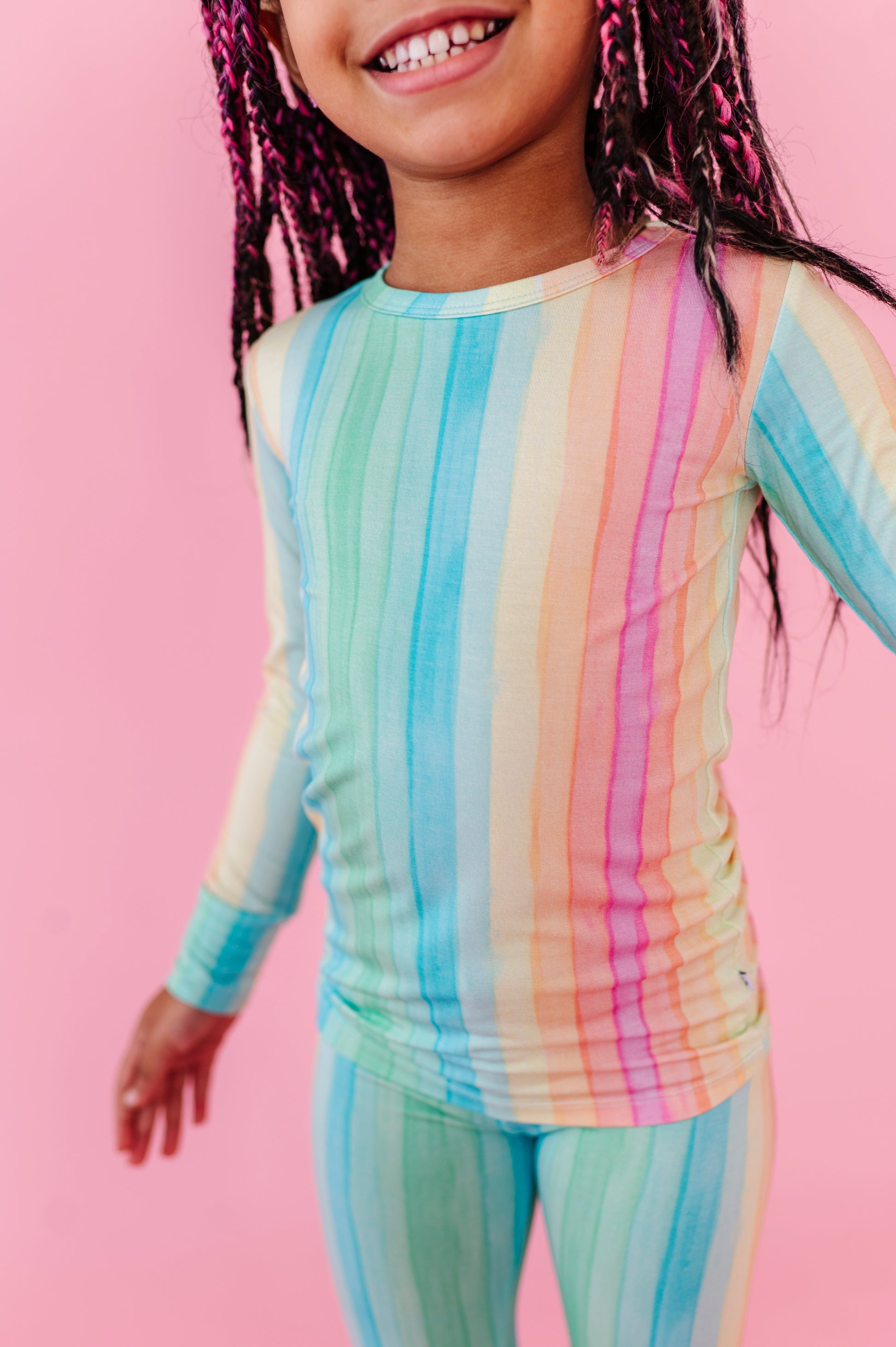 A girl wearing a rainbow striped bamboo pajama two-piece set against a pink background