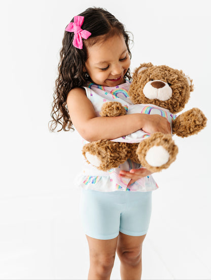 Little girl wearing a bamboo watercolor rainbow peplum top and holding a teddy bear wearing a matching bamboo watercolor rainbow dress 