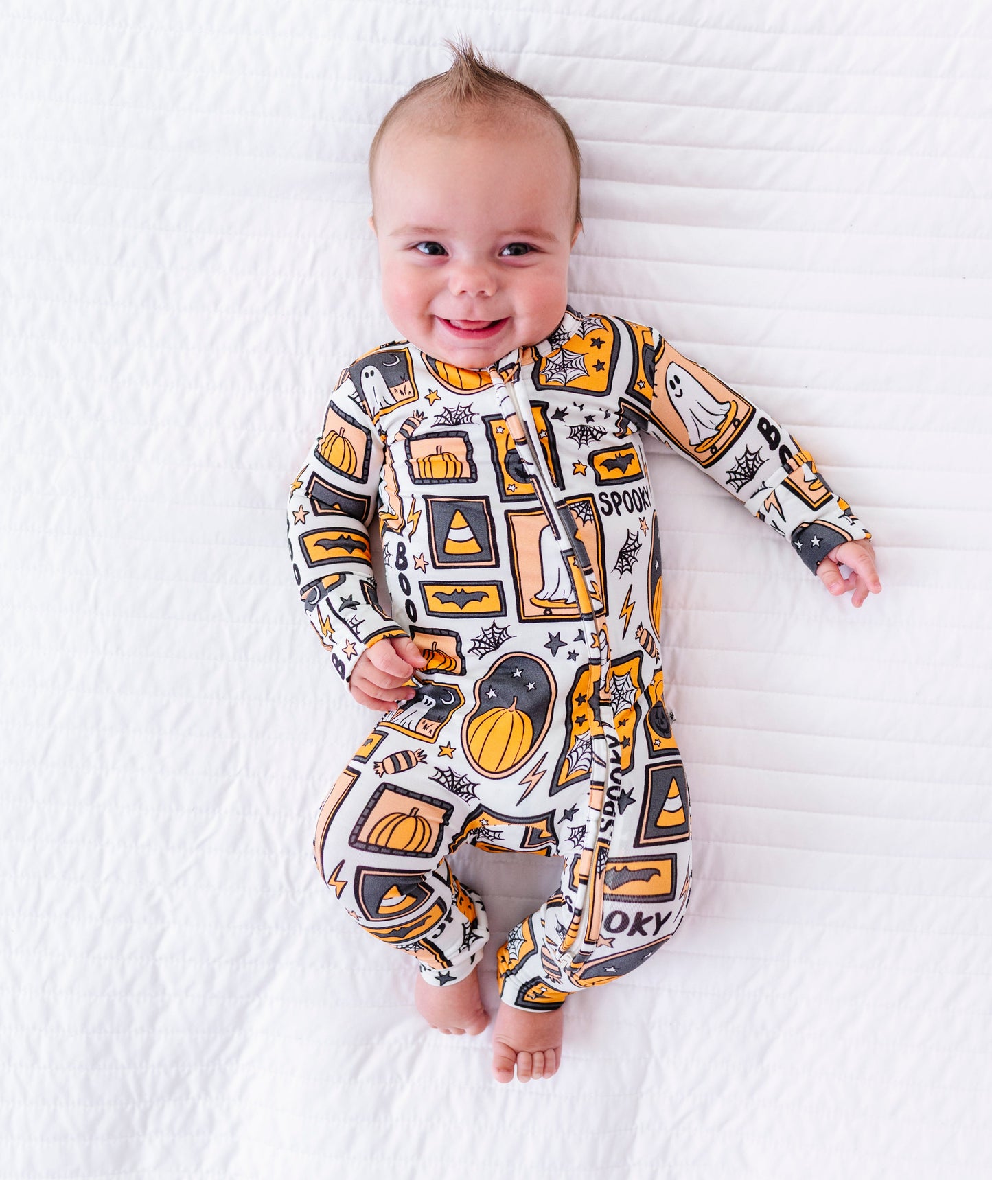 A baby lying on a white textured blanket, wearing a long-sleeve one-piece jumpsuit with a colorful graphic print featuring pumpkins, basketballs, and the word 'JACKSON' in a bold block style.