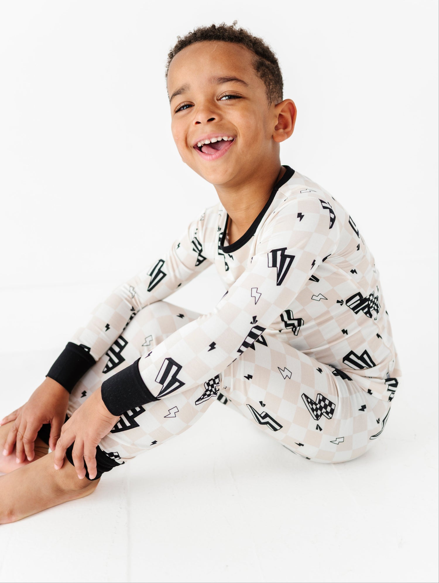 A young boy smiling and laughing while wearing a long-sleeve pajama set with a black and white graphic pattern, sitting against a seamless white background.