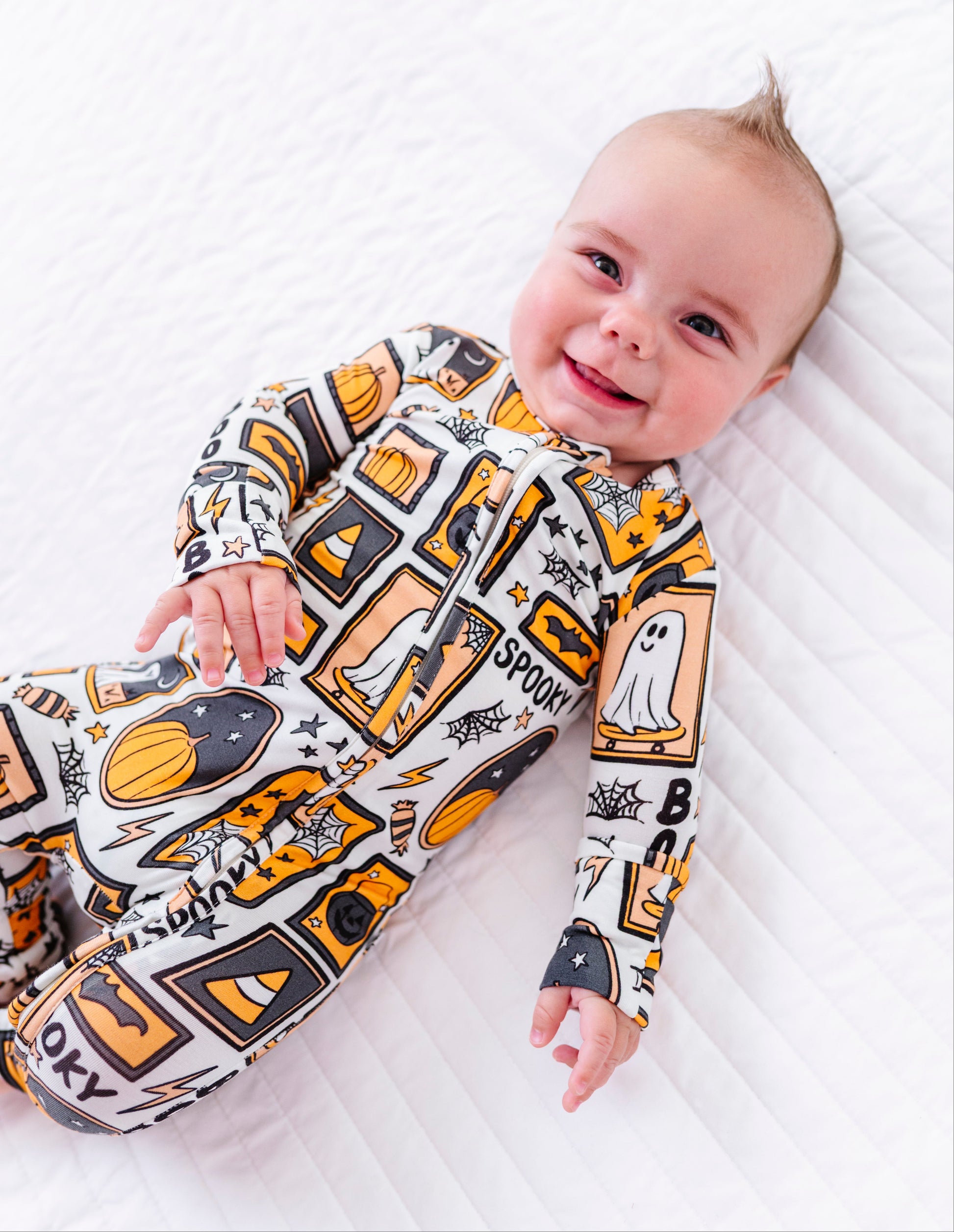 A baby lying on a white quilted mattress, wearing a long-sleeve footed sleeper with a Halloween-themed print featuring ghosts, pumpkins, bats, and the word 'SPOOK'.