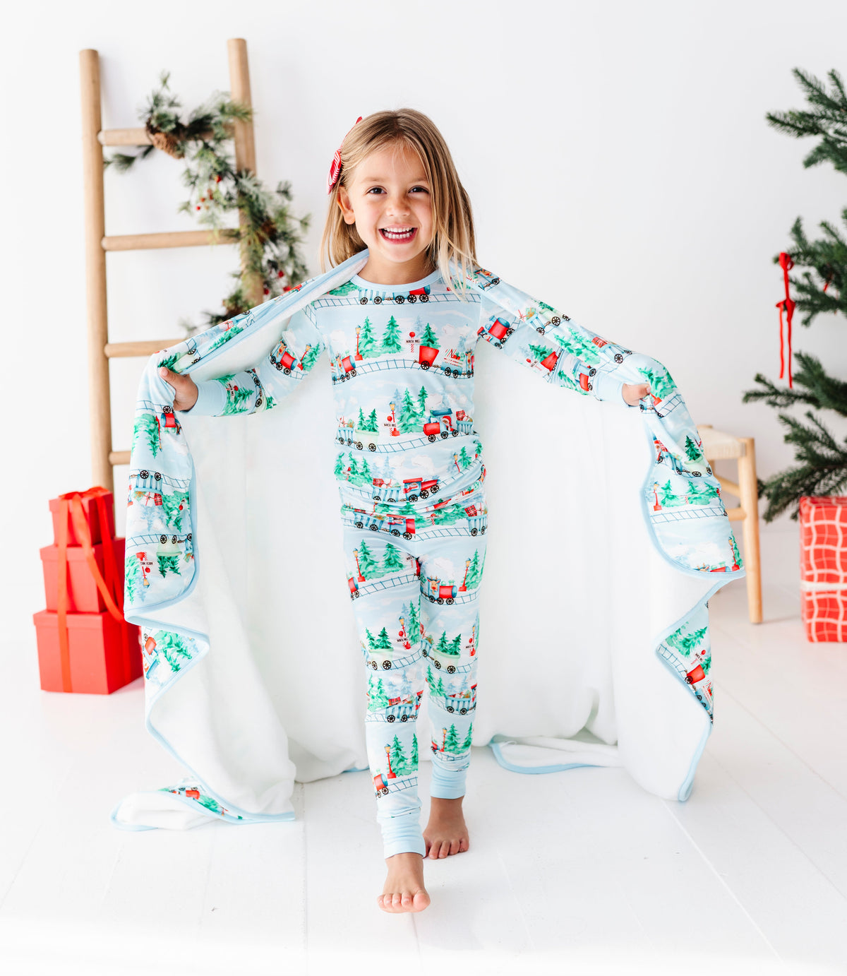 A young girl stands barefoot in a bright white room, holding open a matching holiday-themed bamboo pajama set and blanket. The pajamas and blanket are light blue with a festive Christmas print featuring red trucks, green trees, and white snowflakes.