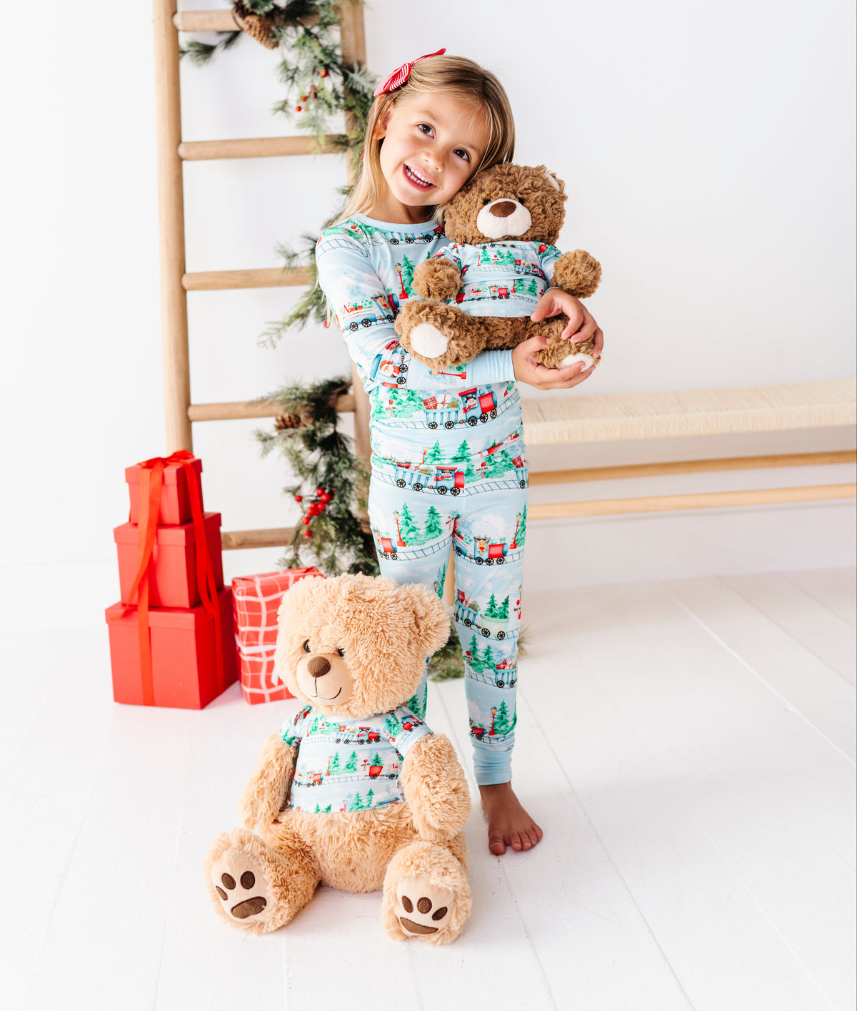 A young girl stands barefoot in a bright white room wearing a light blue Christmas-themed bamboo pajama set with a red and green holiday print. She is holding a brown teddy bear, with a larger teddy bear in front of her and a wooden ladder decorated with greenery in the background.