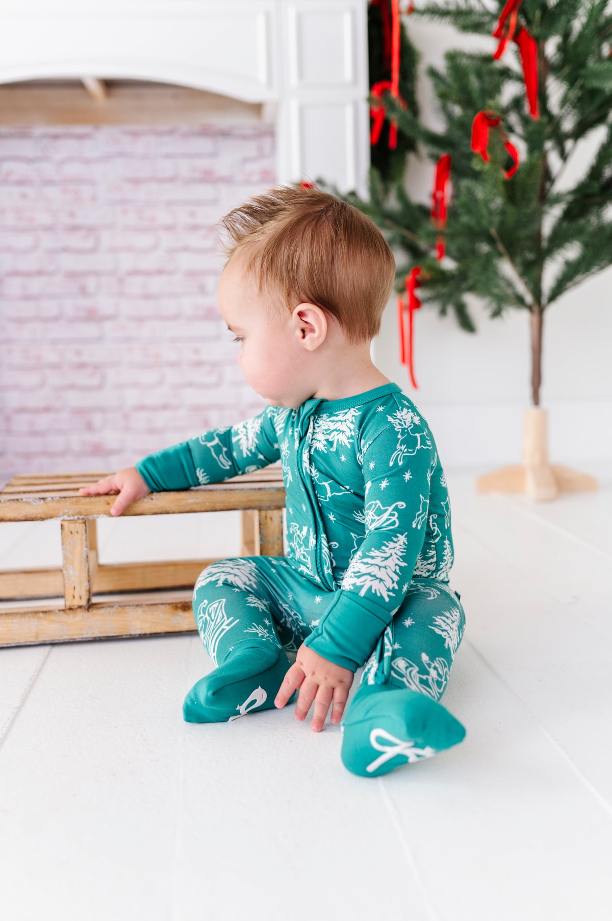 A little boy playing with a wooden sleigh in front of a fireplace. The baby is wearing a bamboo convertible romper featuring vintage Santa and his reindeer 