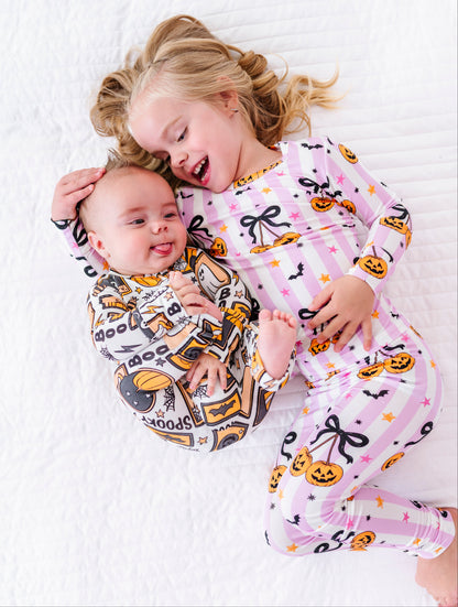 Two children lying on a white quilted bed wearing matching Halloween-themed long-sleeve pajamas. The older child has blonde hair and wears a light pink pajama set with black bows, orange pumpkins, and small stars.