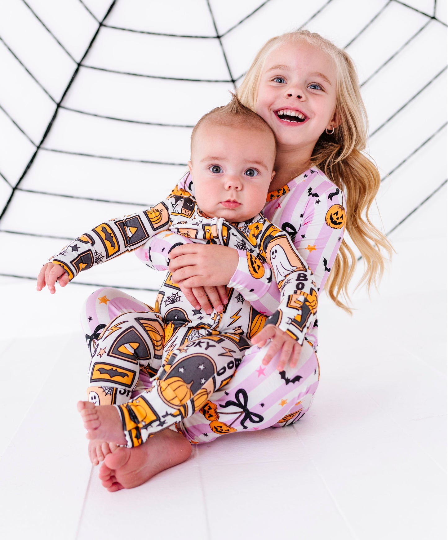 Two children wearing matching pink Halloween-themed long-sleeve pajamas with a spiderweb pattern, sitting in front of a white studio backdrop with a black spiderweb graphic.