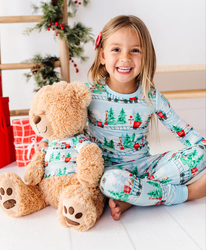 A young girl with blonde hair sits cross-legged on a white wooden floor, smiling and holding a large brown teddy bear. She is wearing a light blue long-sleeve bamboo pajama set with a Christmas tree and Santa Claus print.