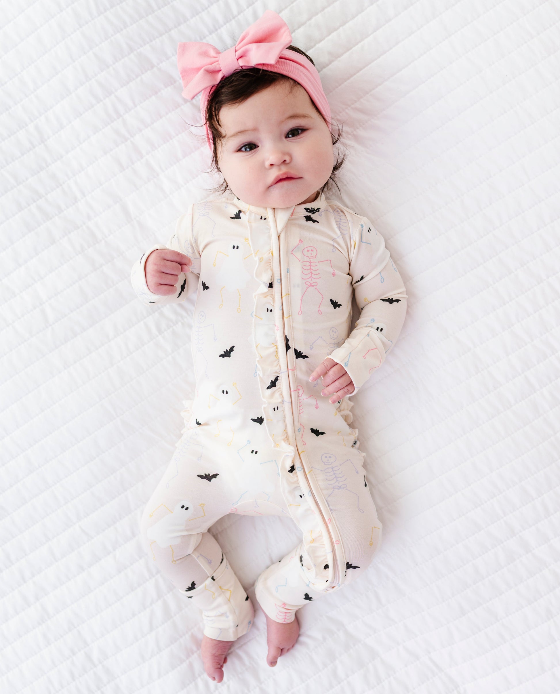 Baby wearing a cream onesie with bat and Halloween  patterns and a pink headband on a white textured surface.