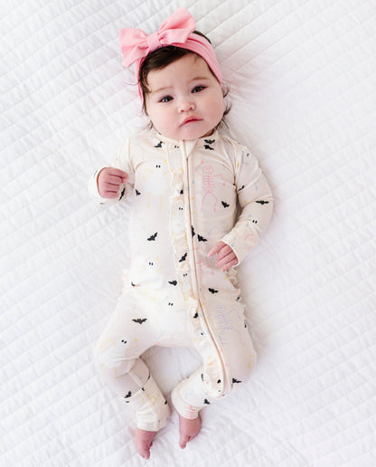 Baby wearing a cream onesie with bat and Halloween  patterns and a pink headband on a white textured surface.