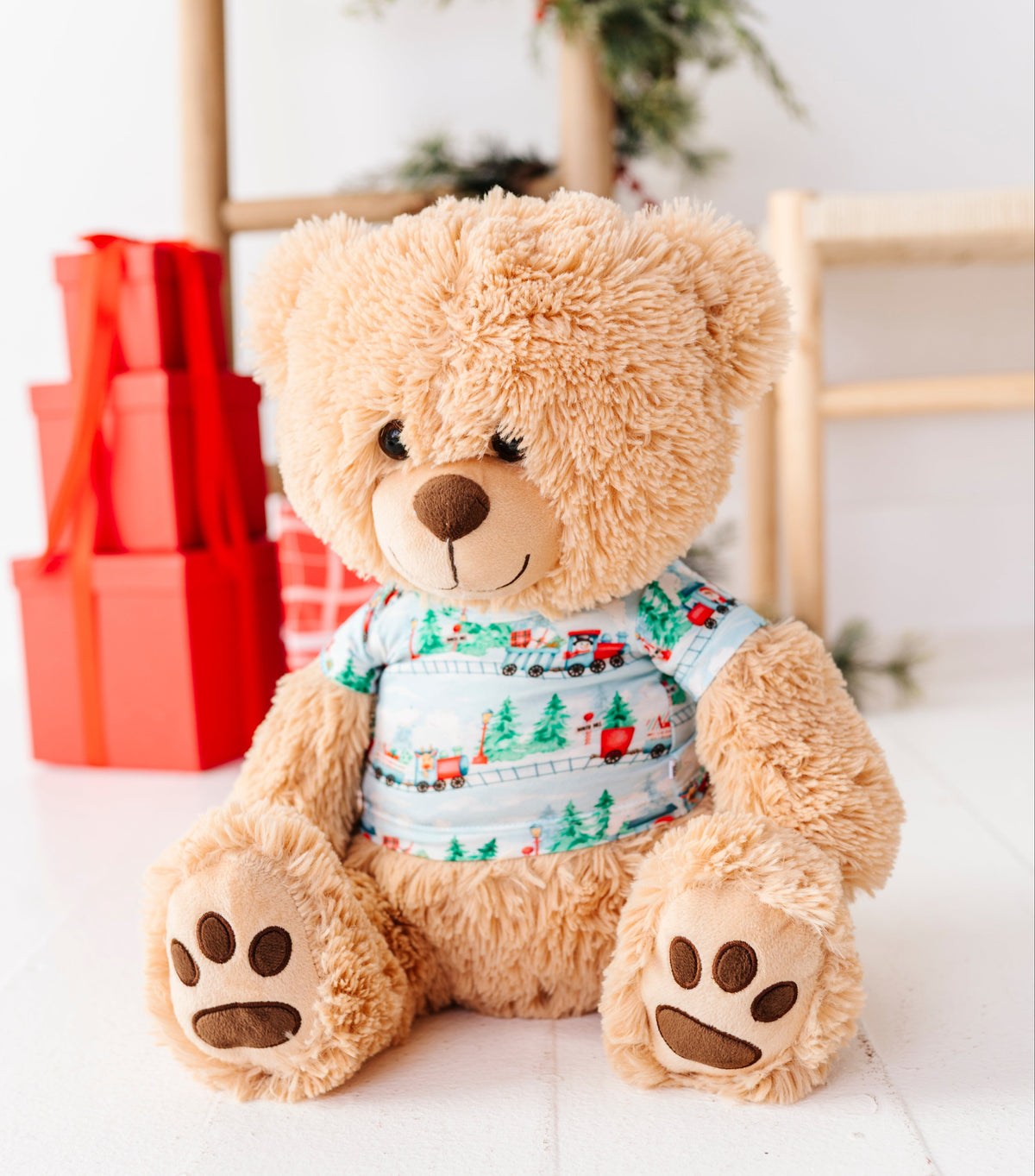 A plush teddy bear in light brown faux fur sits on a white floor, wearing a festive Christmas teddy tee with a light blue background and train print. The background is softly blurred with a wooden ladder, greenery, and red gift boxes.