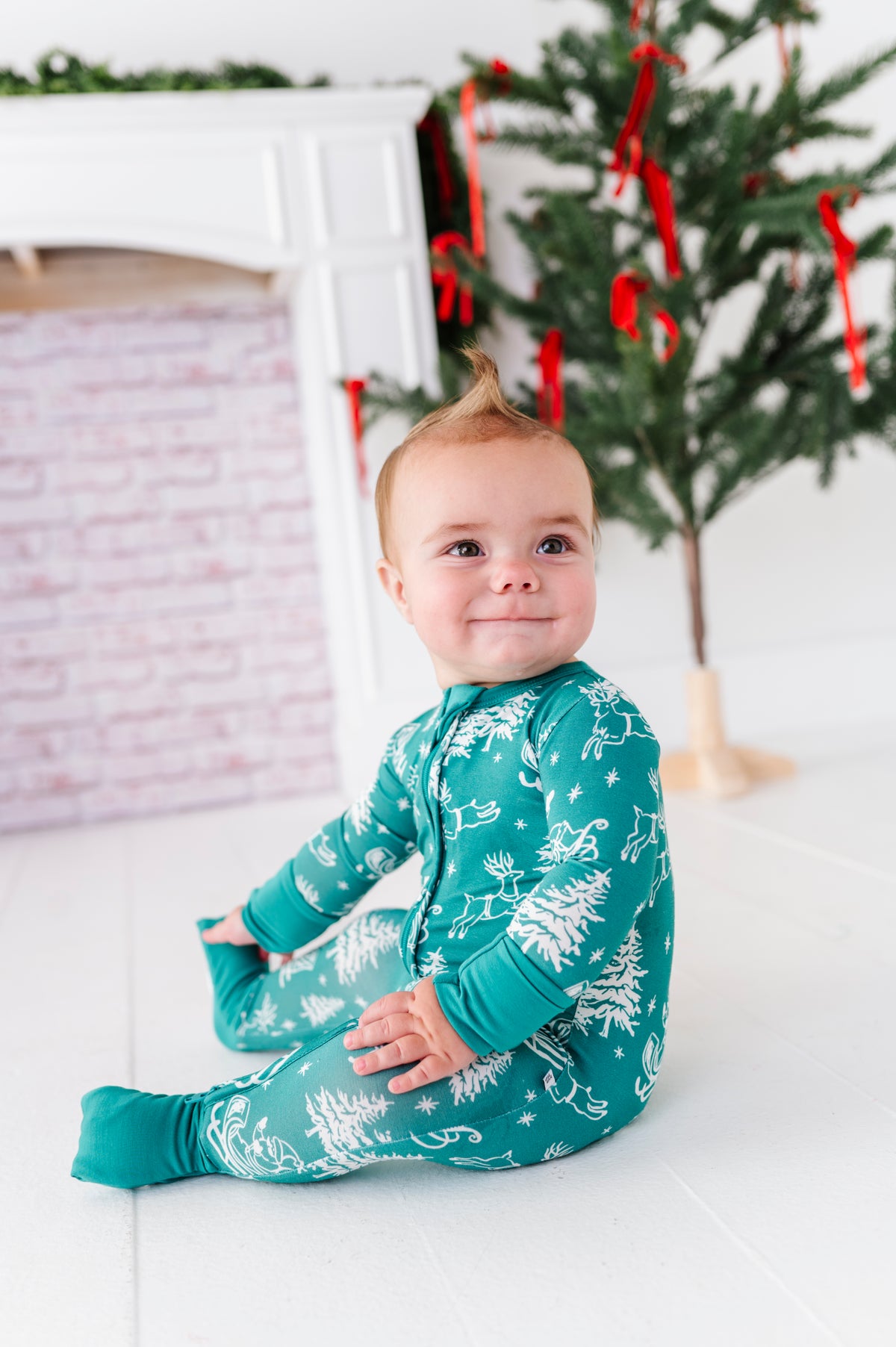 A little boy sitting in front of a fireplace and a Christmas tree wearing a bamboo convertible romper featuring Santa and his reindeer 