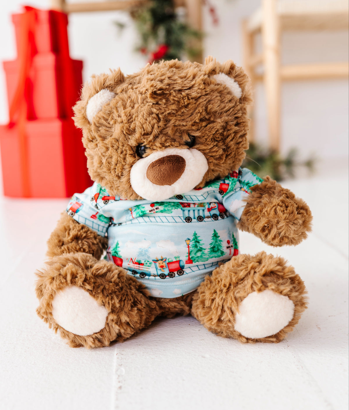 A brown plush teddy bear wearing a festive Christmas-themed bamboo blue shirt with red and green train and tree patterns, sitting on a white wooden surface with blurred holiday decorations in the background.