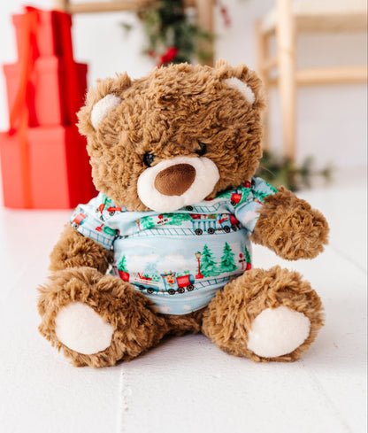 A brown plush teddy bear wearing a festive Christmas-themed bamboo blue shirt with red and green train and tree patterns, sitting on a white wooden surface with blurred holiday decorations in the background.