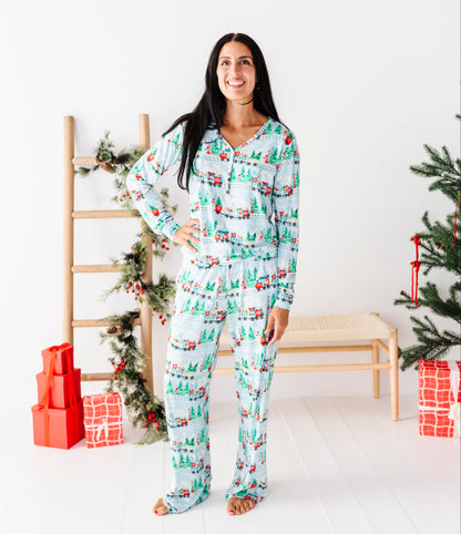 A woman stands in a bright white room wearing a long-sleeve pajama set with a Christmas tree and snowman print. The pajamas are light blue with green trees and red accents, paired with matching pants.
