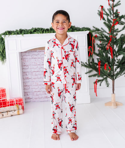 A young boy wearing a loose-fitting bamboo lounge set featuring Santa throwing baseballs and knocking them out of the park! He's standing in front of a fireplace and a blurry Christmas tree