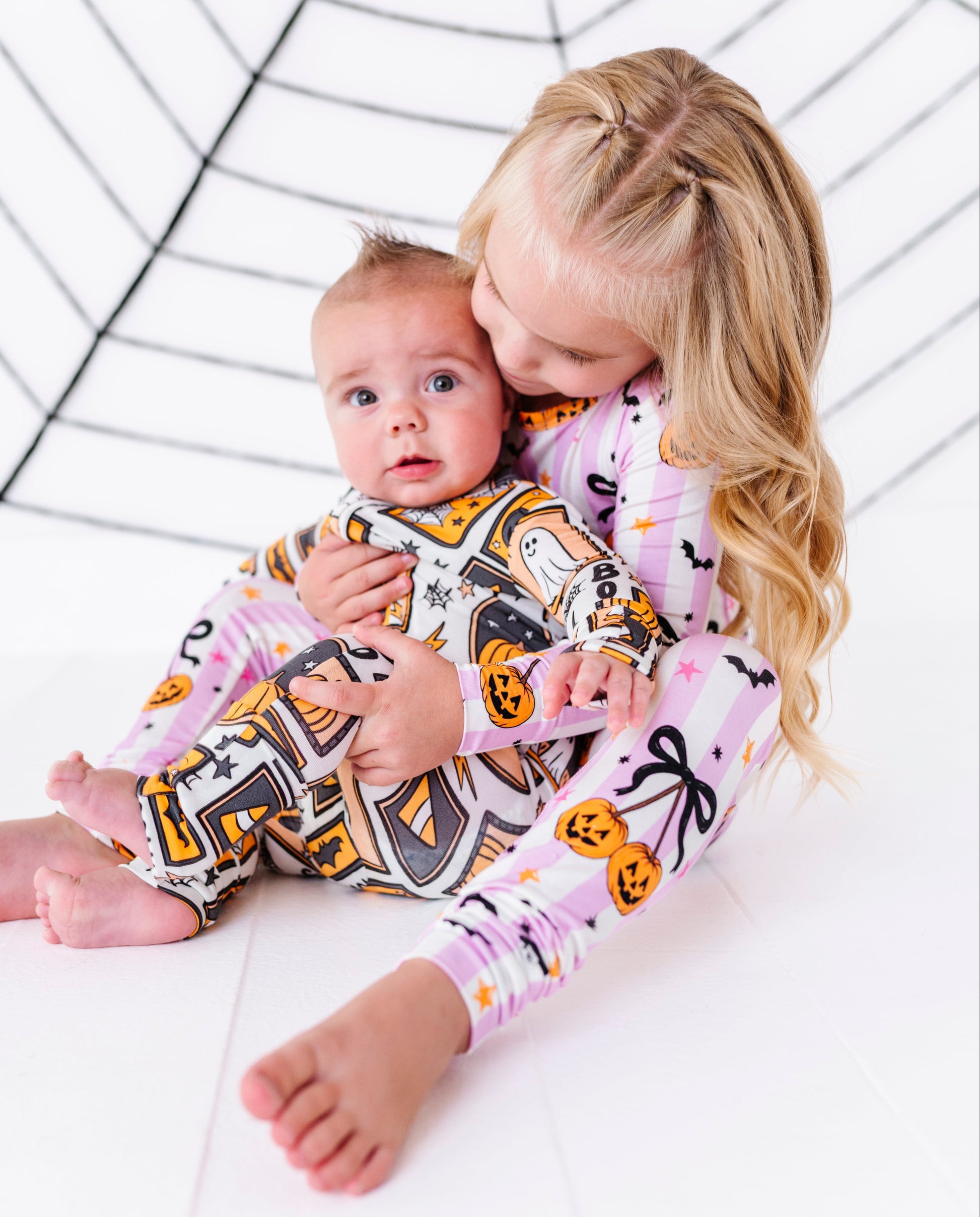 A young girl with blonde hair and a baby sitting together on a white floor, both wearing matching pink and white Halloween-themed patterned pajamas with black spiderwebs and orange jack-o'-lanterns, against a white background with a black spiderweb graphic.