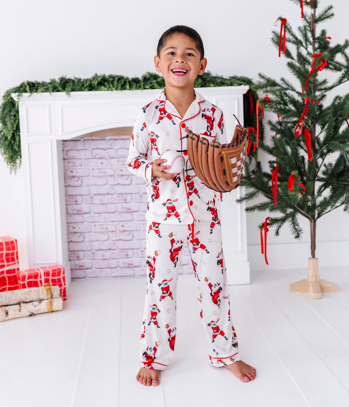 A little boy wearing a loose-fitting bamboo lounge set featuring Santa throwing baseballs and knocking them out of the park! He's holding a baseball wearing a leather glove while standing in front of a fireplace and a blurry Christmas tree
