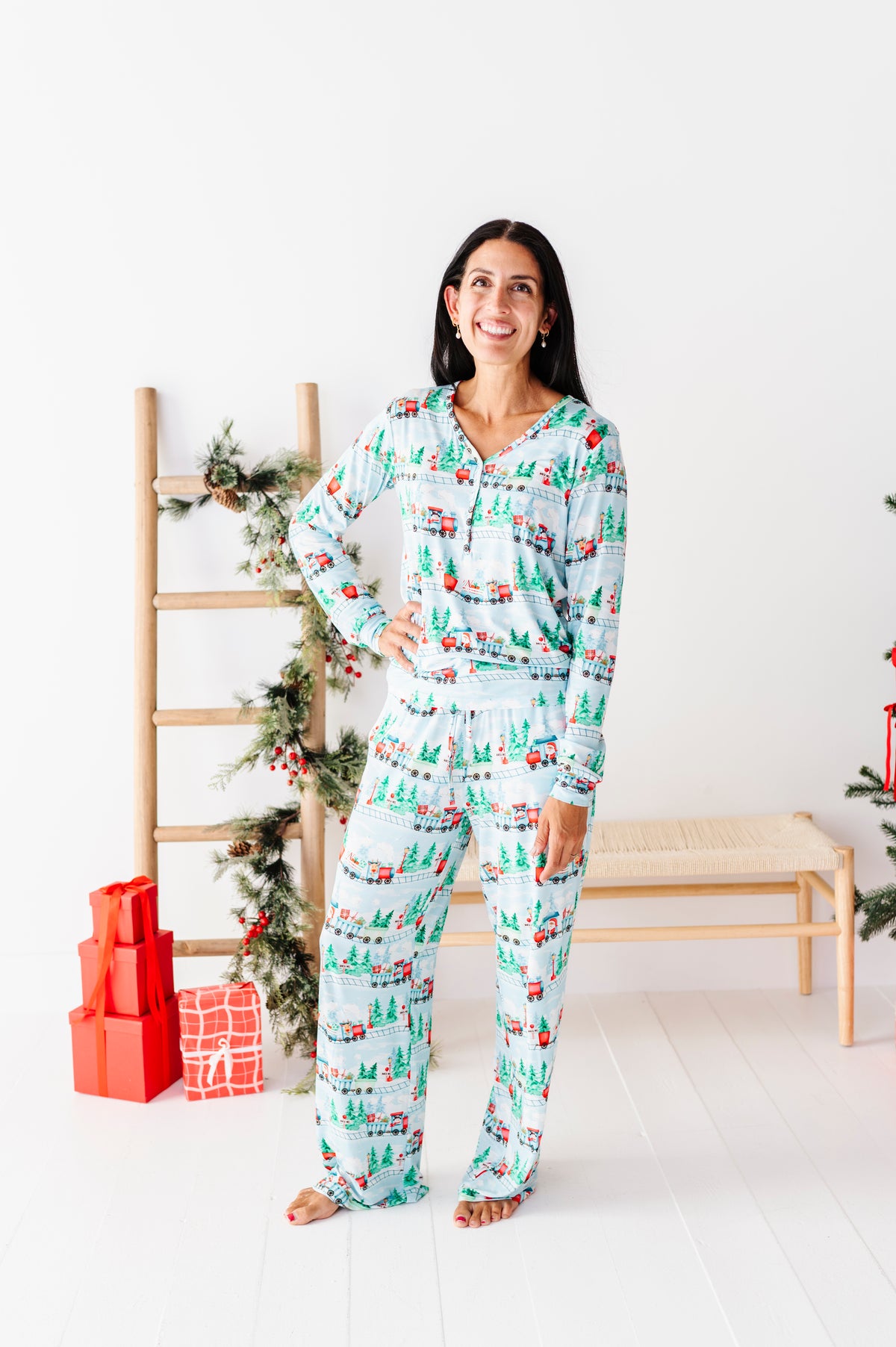 A woman stands barefoot in a bright white room wearing a long-sleeve pajama set with a Christmas tree and gift print. A wooden ladder with holiday garland and wrapped presents is behind her.
