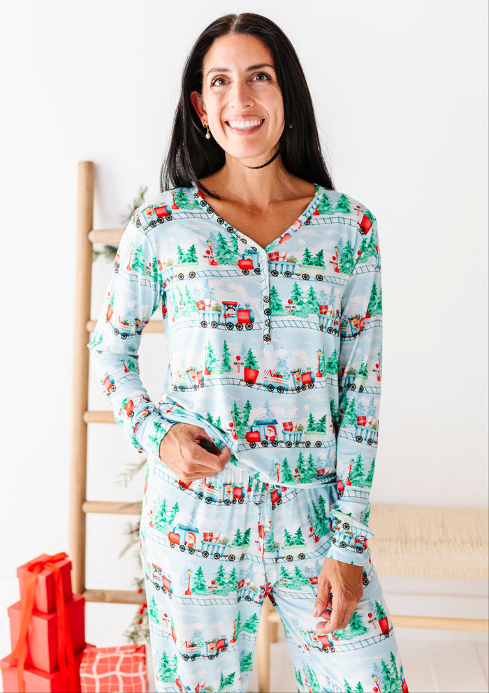 A woman wearing a light blue long-sleeve pajama set with a Christmas village print featuring red trucks, green trees, and small houses, standing in a brightly lit room with a wooden ladder and wrapped gifts.
