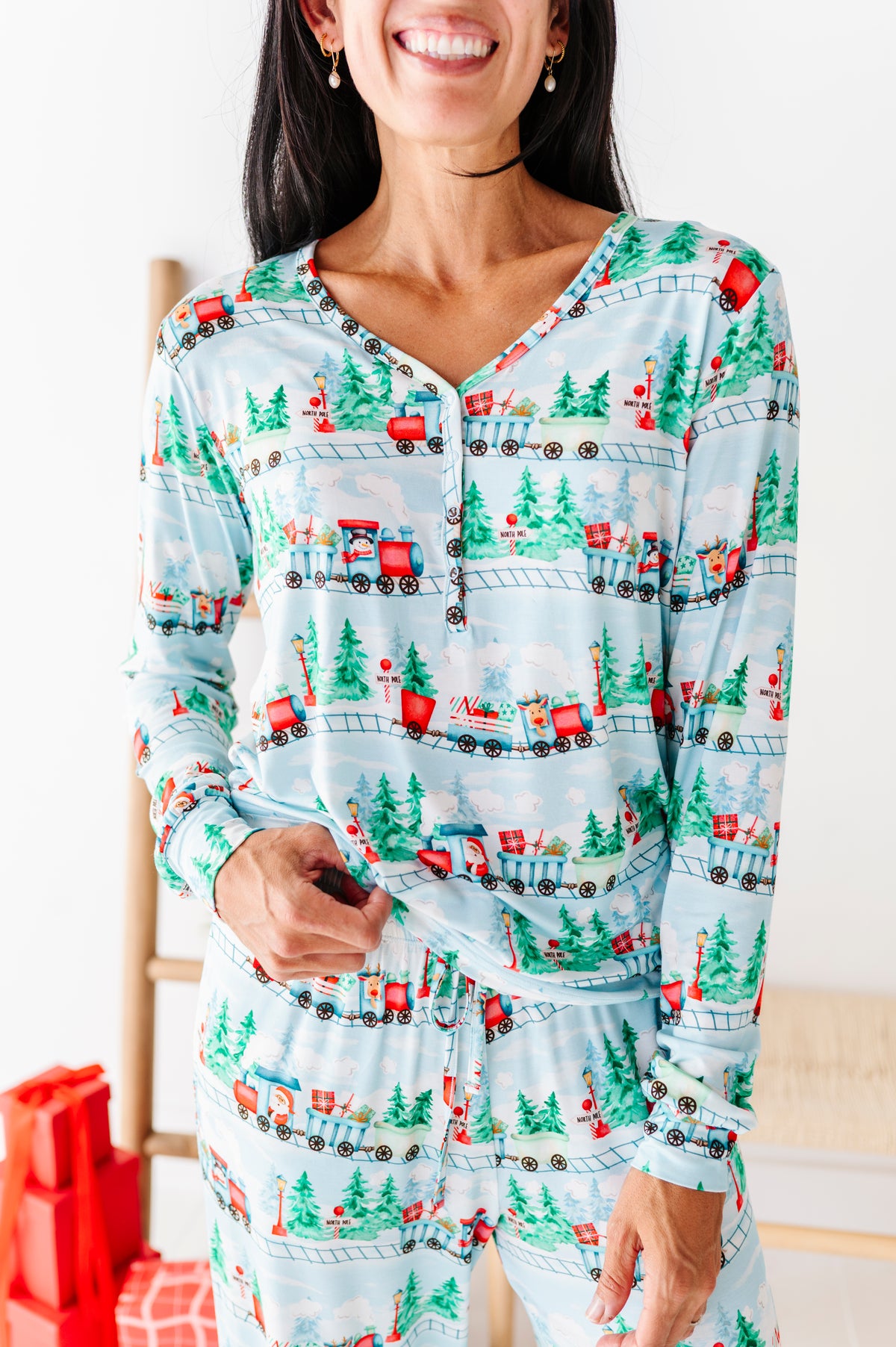 A woman wearing a light blue long-sleeve pajama set with a Christmas train and tree pattern, standing indoors against a white wall.