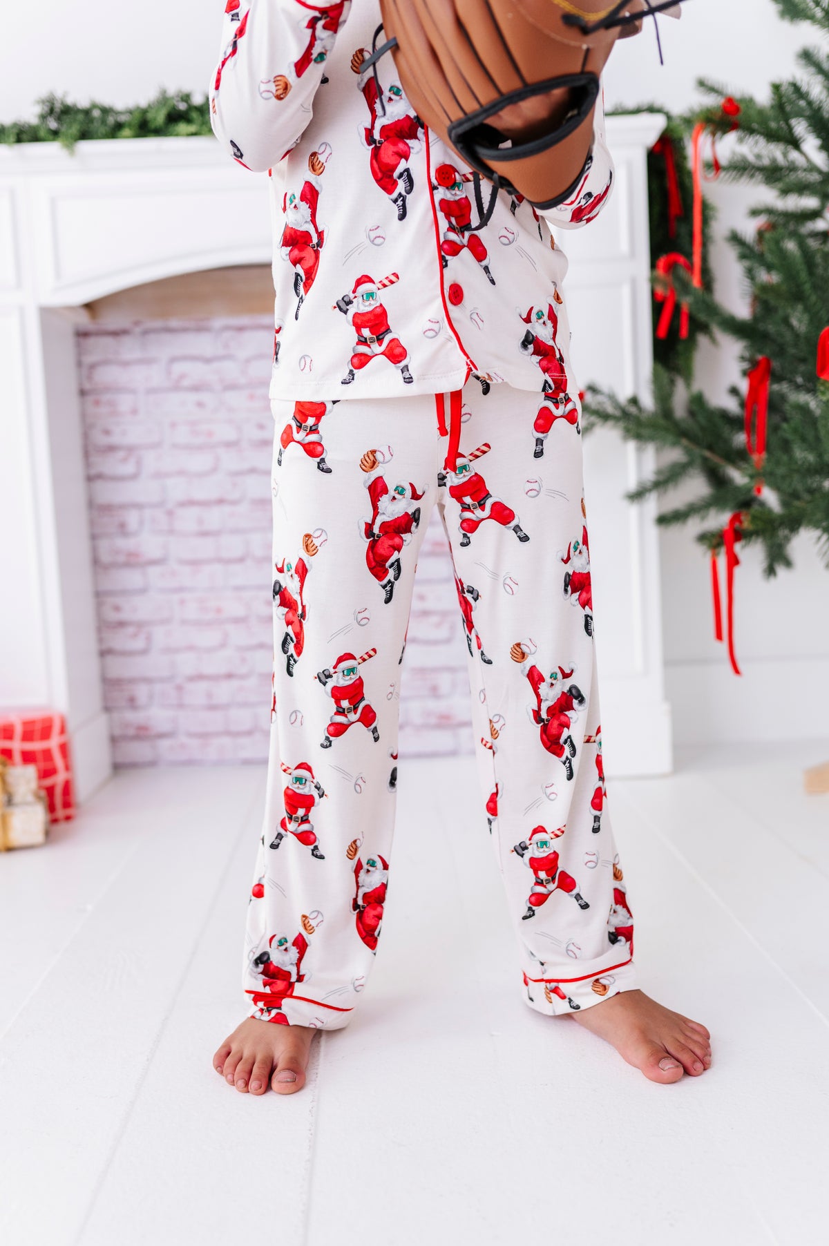 A photo of a little boy wearing a loose-fitting bamboo lounge set featuring Santa throwing baseballs and knocking them out of the park! He's holding a wooden bat while standing in front of a fireplace and a blurry Christmas tree