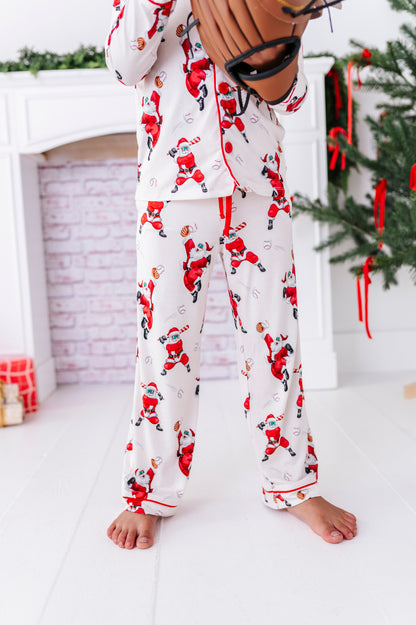 A photo of a little boy wearing a loose-fitting bamboo lounge set featuring Santa throwing baseballs and knocking them out of the park! He's holding a wooden bat while standing in front of a fireplace and a blurry Christmas tree