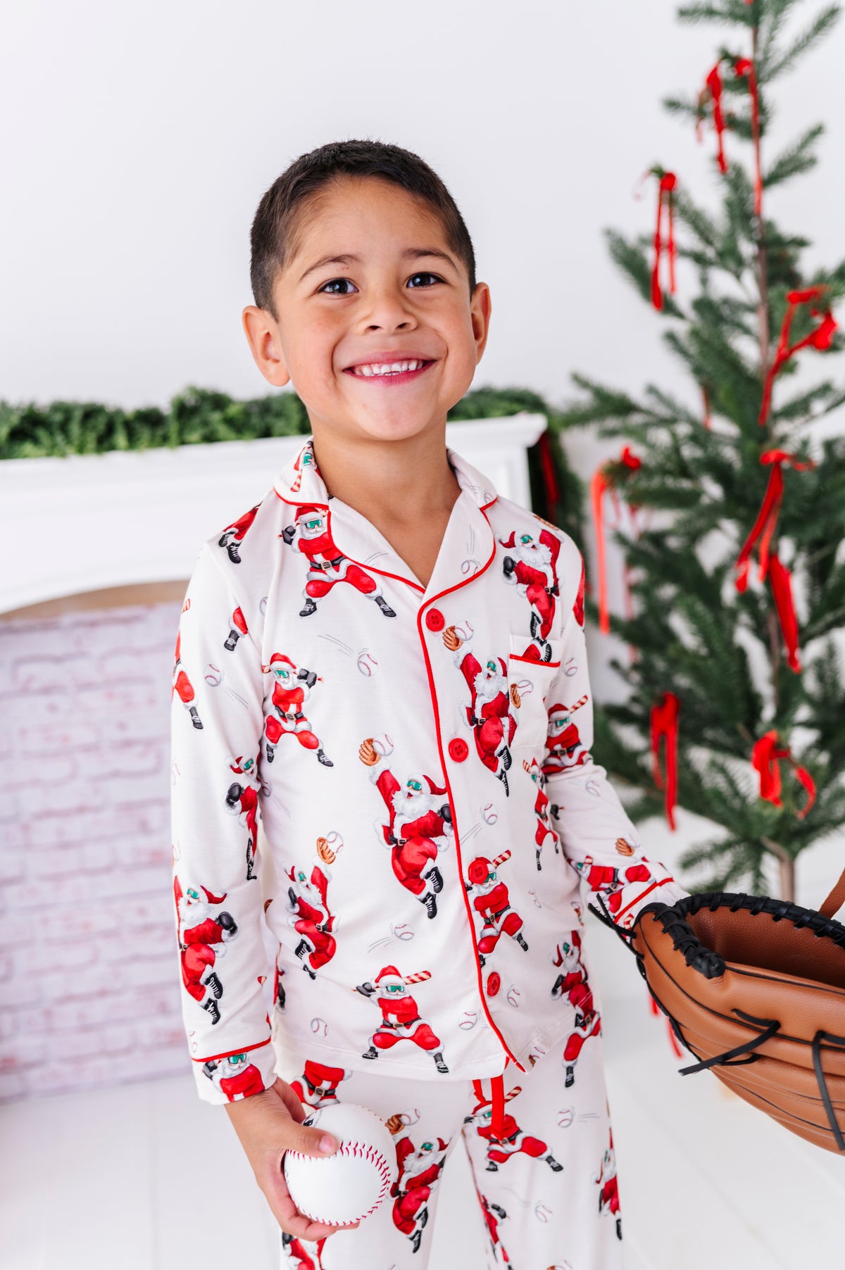 A little boy wearing a loose-fitting bamboo lounge set featuring Santa throwing baseballs and knocking them out of the park! He's holding a wooden bat while standing in front of a fireplace and a blurry Christmas tree