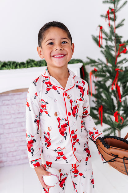 A little boy wearing a loose-fitting bamboo lounge set featuring Santa throwing baseballs and knocking them out of the park! He's holding a wooden bat while standing in front of a fireplace and a blurry Christmas tree