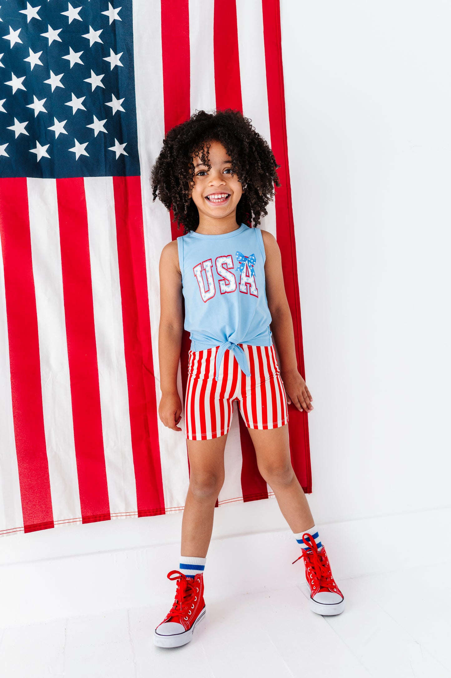 Child wearing a 'USA' tank top and red and white striped bamboo biker shorts standing in front of an American flag.