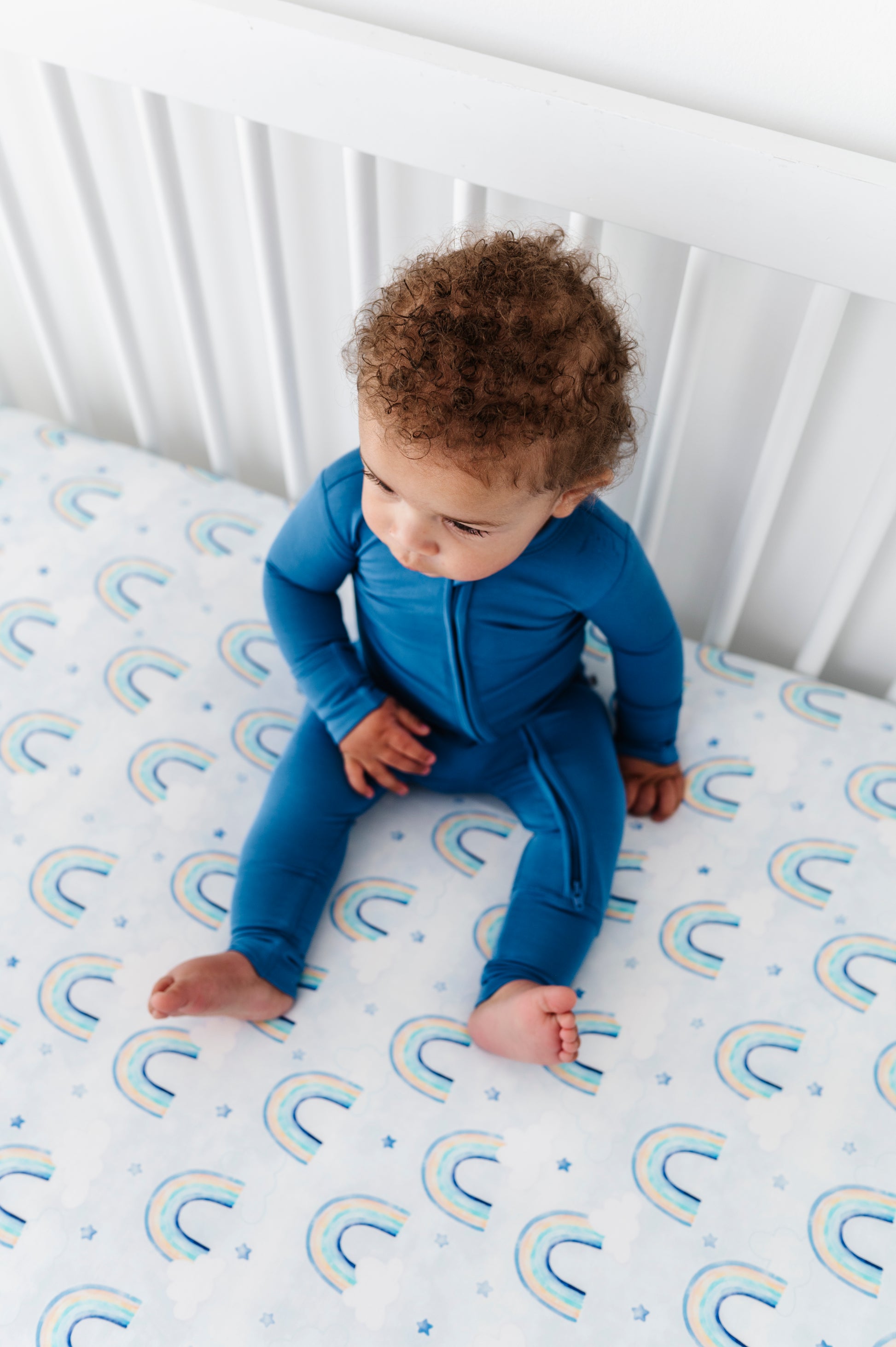 Child in a blue bamboo zip romper sitting on a crib with a rainbow-patterned bamboo crib sheet