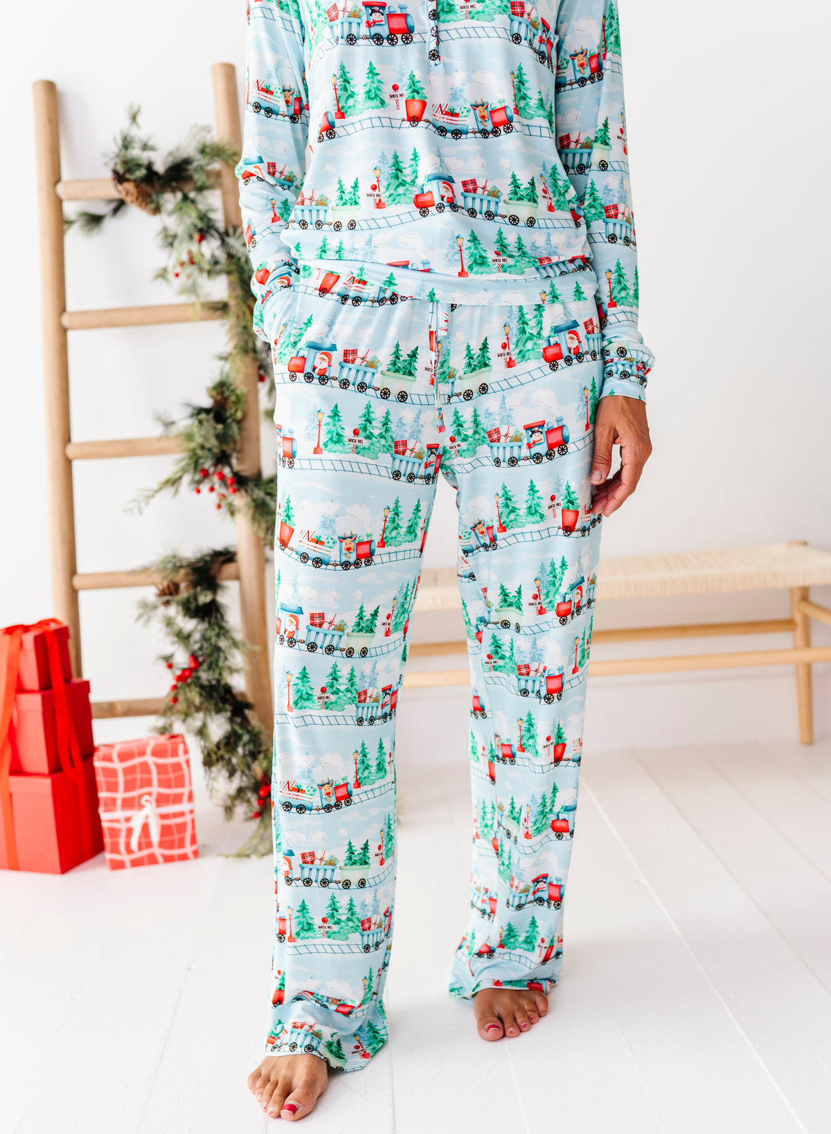 A person wearing a light blue long-sleeve pajama set with a Christmas village print featuring green trees, red houses, and small vehicles, standing barefoot in a bright room with holiday decor and wrapped gifts in the background.