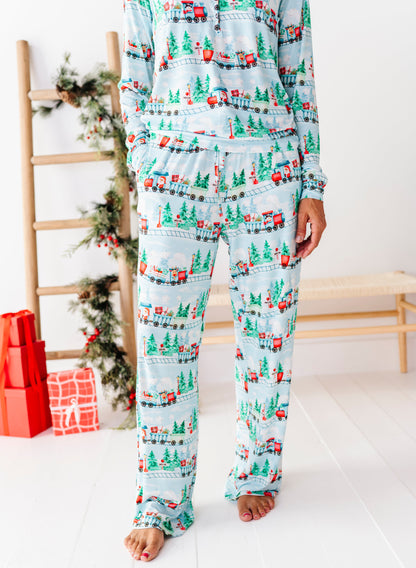 A person wearing a light blue long-sleeve pajama set with a Christmas village print featuring green trees, red houses, and small vehicles, standing barefoot in a bright room with holiday decor and wrapped gifts in the background.