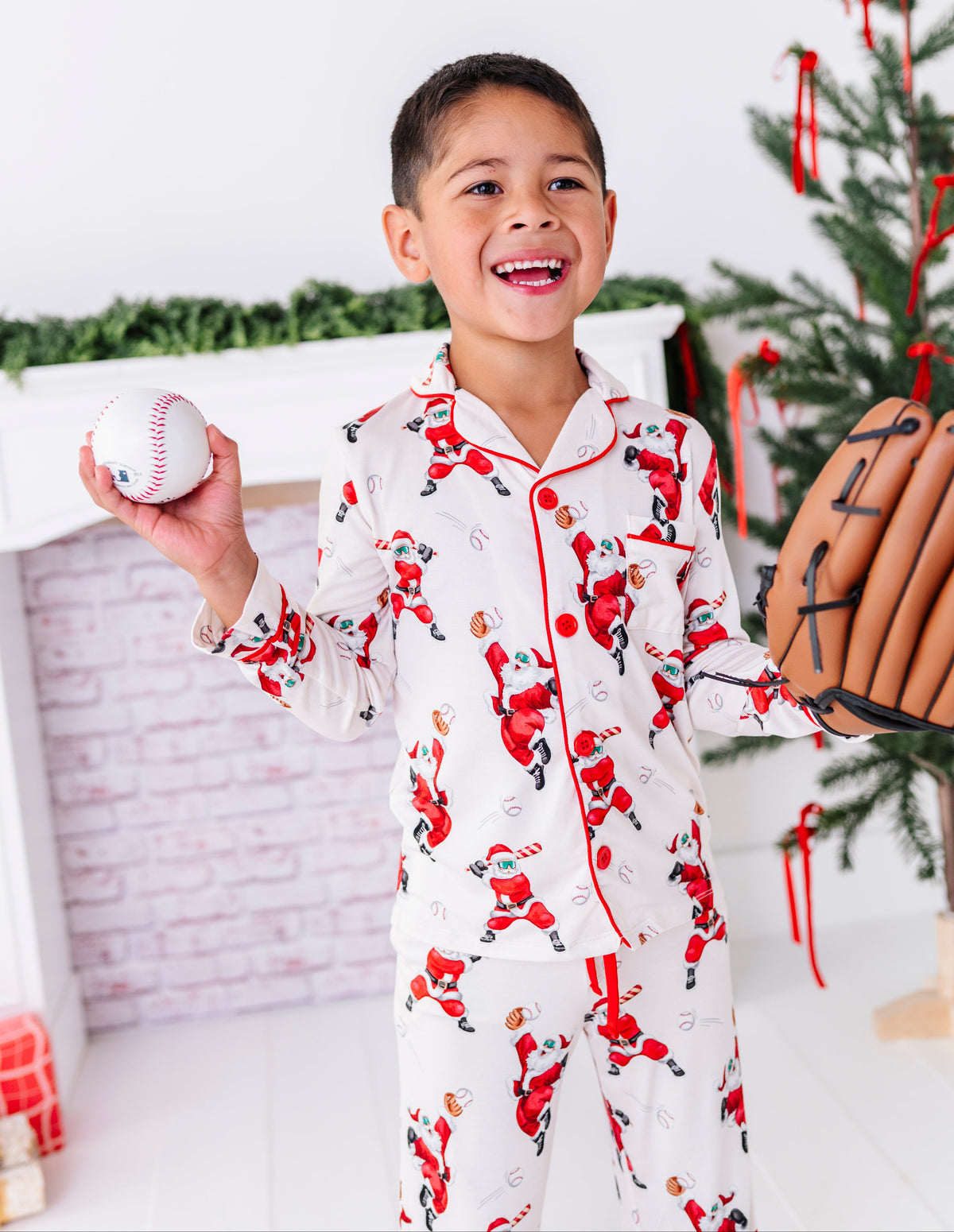 A little boy wearing a bamboo lounge set featuring Santa throwing baseballs and knocking them out of the park! He's holding a wooden bat while standing in front of a fireplace and a blurry Christmas tree