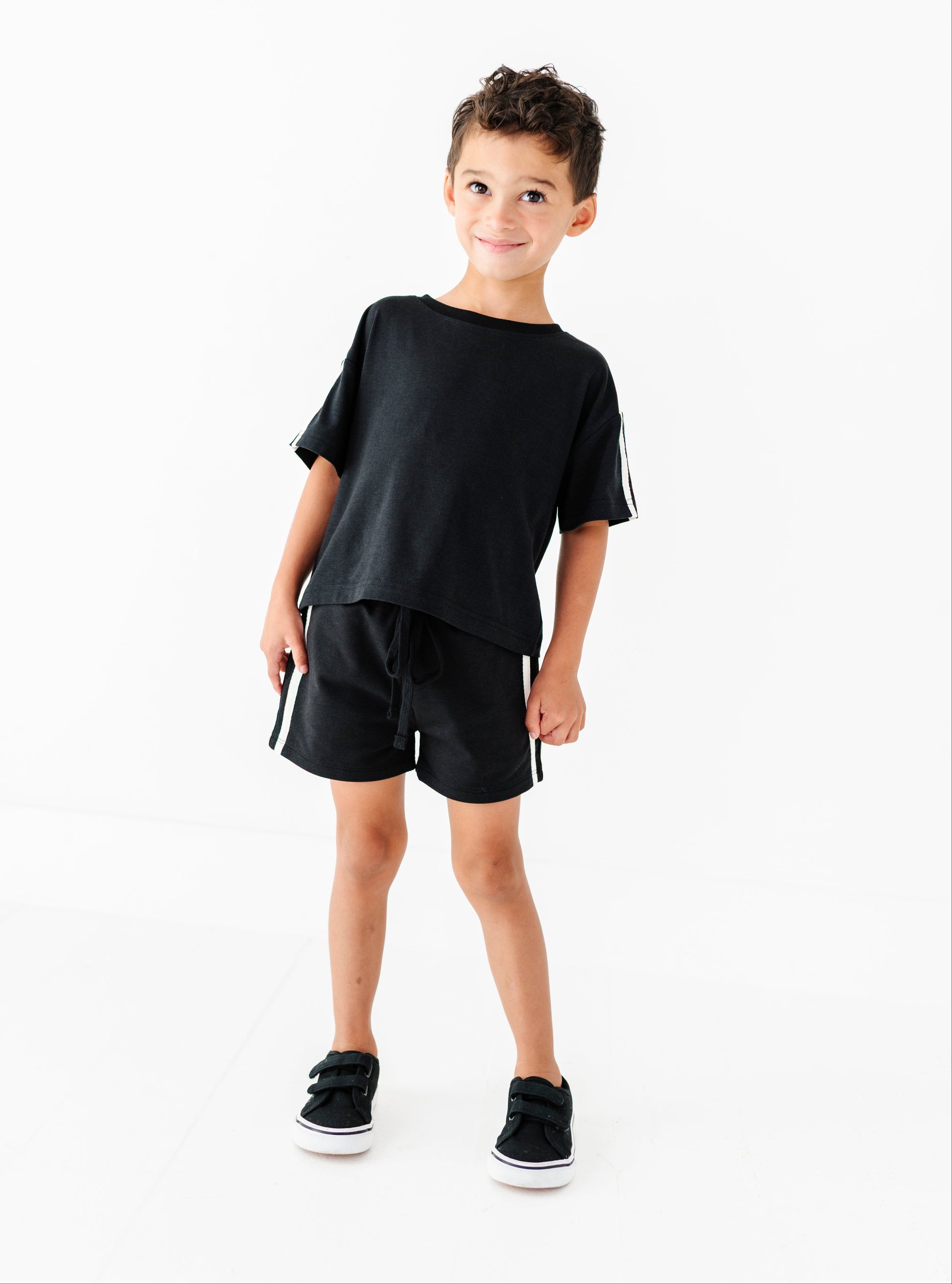 A young boy modeling a black short-sleeve t-shirt and matching black shorts with white side stripes, paired with black sneakers, against a solid white studio background.