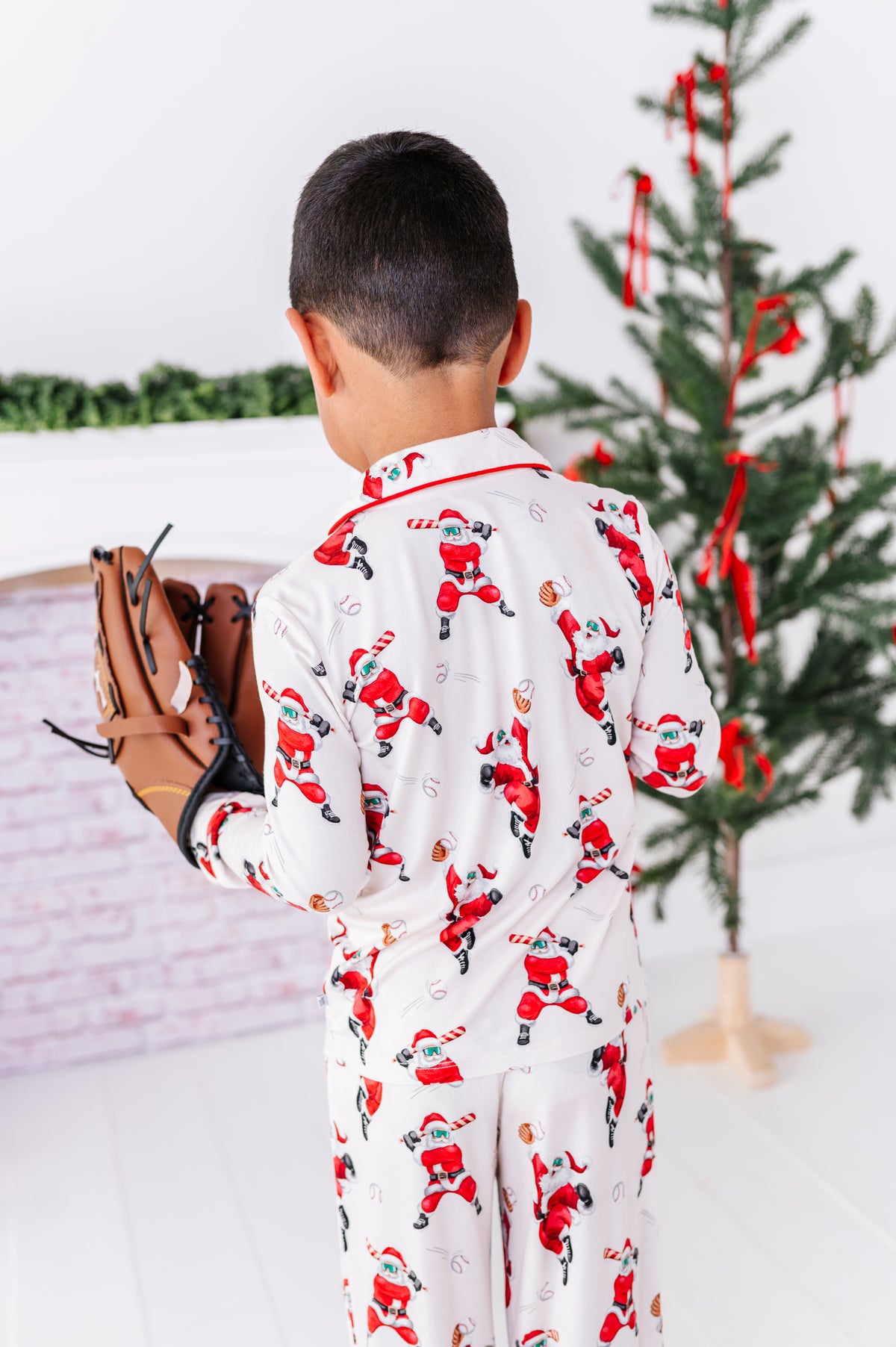A little boy wearing a loose-fitting bamboo lounge set featuring Santa throwing baseballs and knocking them out of the park! He's standing with his back to the camera wearing a leather glove while standing in front of a fireplace and a blurry Christmas tree