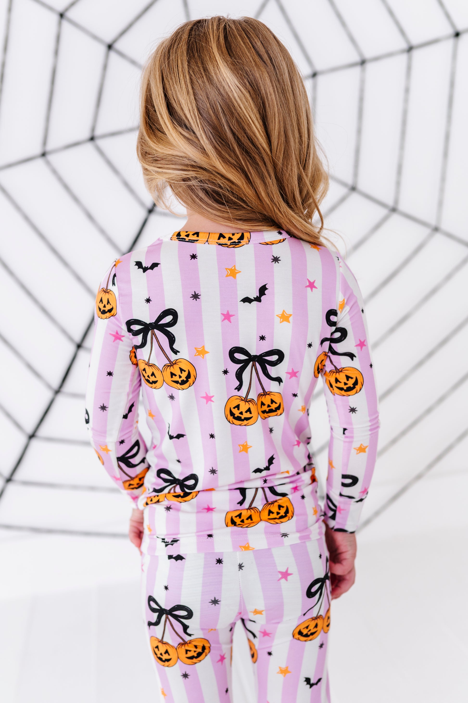 A child viewed from behind wearing a pink and white striped pajama set with a Halloween print of pumpkins, black bows, bats, and stars, set against a white background with a black spiderweb graphic.