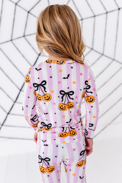 A child viewed from behind wearing a pink and white striped pajama set with a Halloween print of pumpkins, black bows, bats, and stars, set against a white background with a black spiderweb graphic.