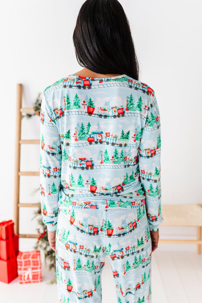 A person viewed from behind wearing a matching long-sleeve pajama set with a Christmas train print on a light blue background, standing indoors near a wooden ladder and wrapped gifts.