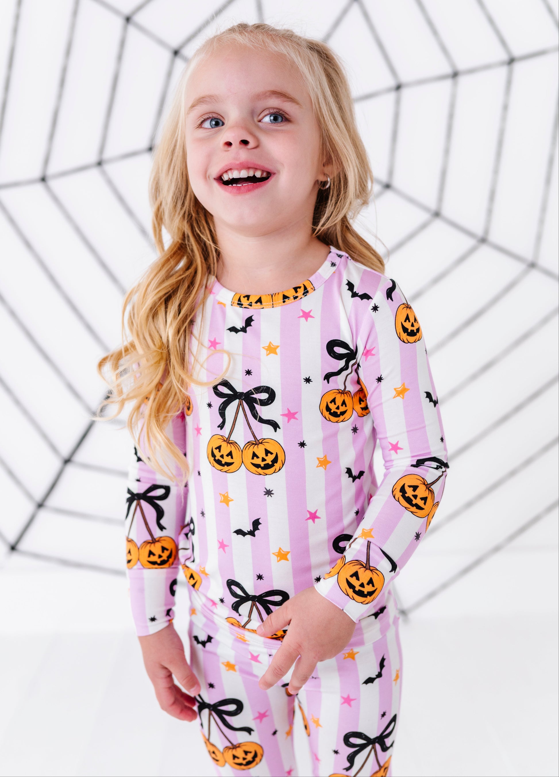 A young girl with blonde hair smiling and looking to the side, wearing a pink and white striped long-sleeve pajama set with a Halloween print of pumpkins, black bows, and stars. She is standing in front of a white background with a black spiderweb graphic.