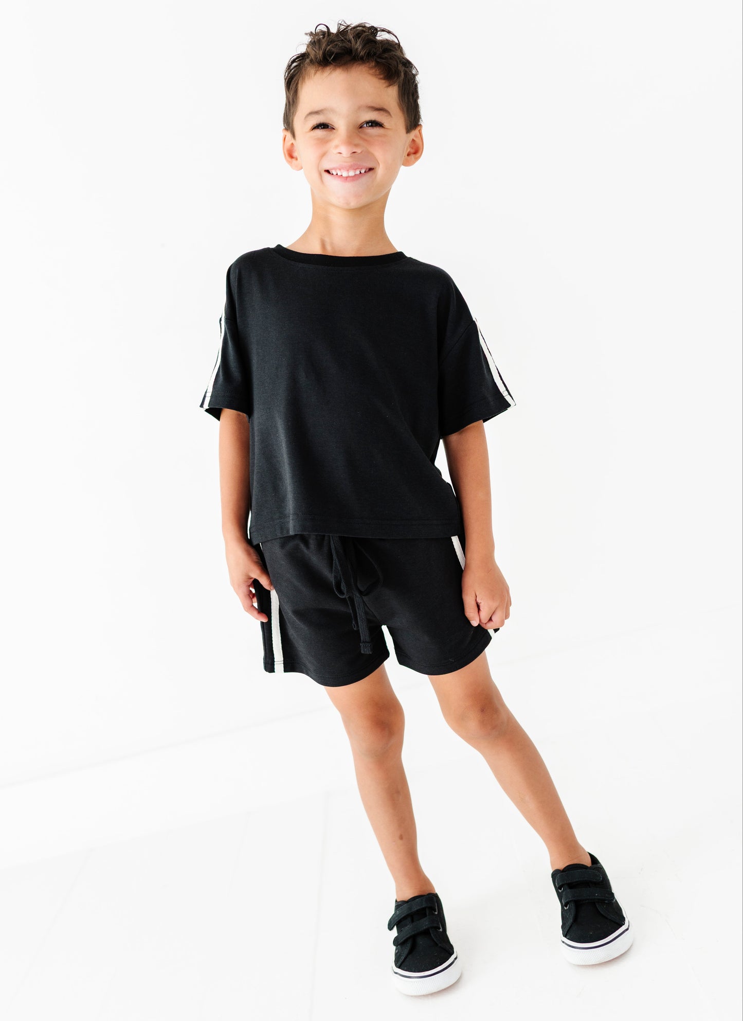 A young boy modeling a black short-sleeve t-shirt and matching black shorts with white side stripes, paired with black sneakers, against a clean white background.